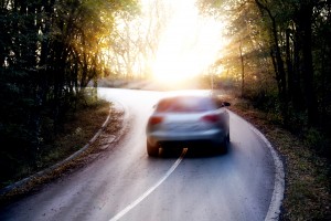 image of car speeding around bend on country road with sun shining through trees.